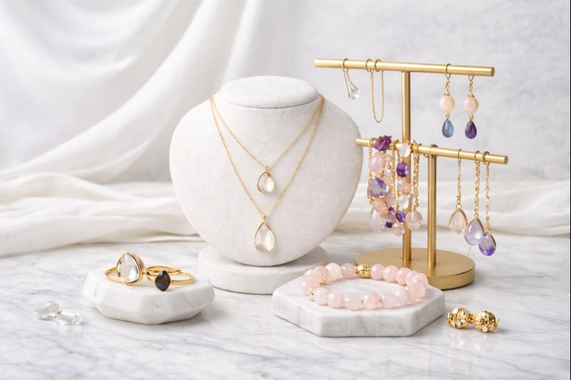 “Elegant display of gemstone jewelry including gold necklaces, bracelets, rings, and earrings arranged on white marble with soft draped fabric, featuring clear quartz, amethyst, moonstone, and rose quartz.”